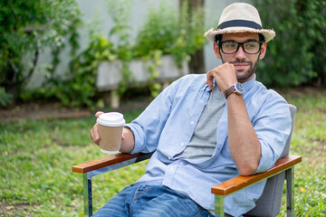 young handsome beard man freelancer sitting on chair holding paper cup of coffee at front yard...