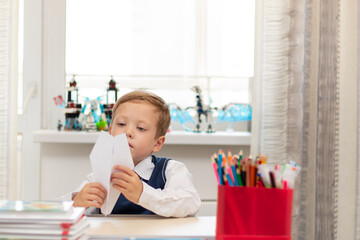 A cute first-grader boy in a school uniform at home while isolated at his desk makes a paper airplane during recess. Selective focus. Portrait