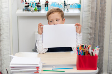 A cute boy first grader in a school uniform at home while isolated at his desk holds a sheet of...