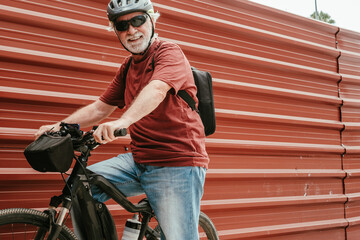 Senior cyclist in outdoor excursion with electric bicycle resting against a red metal panel looking at camera