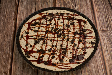 Pay de queso crema decorado con trozos de galleta de chocolate amargo y rayas de chocolate líquido sobre una superficie de madera
