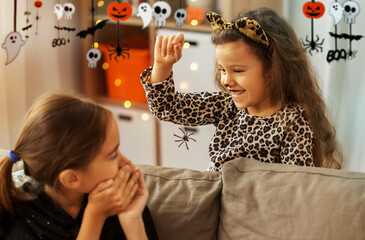 halloween, holiday and childhood concept - smiling little girls in party costumes playing with toy spider at home decorated with garland and lights
