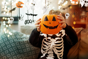 Naklejka premium halloween, holiday and childhood concept - smiling little boy in costume of skeleton with jack-o-lantern sitting on sofa at home