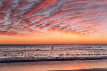 Surfer in the ocean under colourful stripy cloud sunrise skies