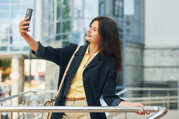 Holding phone in hand. Woman in formal wear standing outdoors in the city at daytime