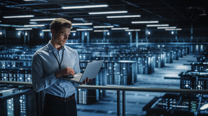 Portrait of IT Specialist Uses Laptop in Data Center. Server Farm Cloud Computing Facility with...