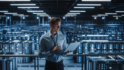 Portrait of IT Specialist Uses Laptop in Data Center. Server Farm Cloud Computing Facility with Male Maintenance Administrator Working on Cyber Security and Network Protection.