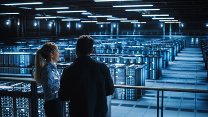 Data Center Male It Specialist and Female e-Commerce Manager Talk, Use Laptop. Big Server Farm Cloud Computing Facility with Two Information Technology Professionals working.