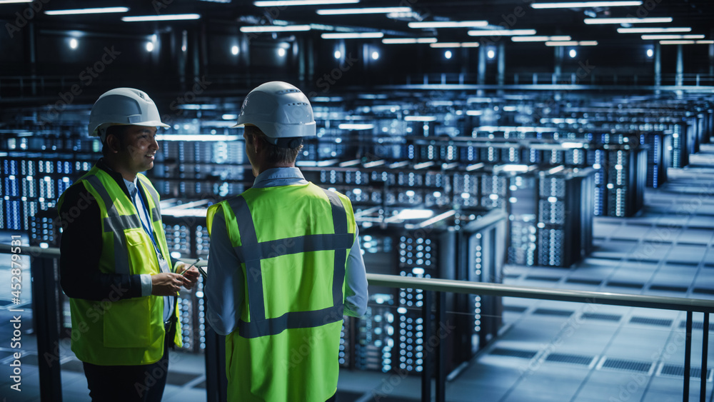 Poster Data Center IT Specialist and System administrator Talk, Use Tablet Computer, Wearing Safety Wests. Server Clod Farm with Two Information Technology Engineers checking Cyber Security.