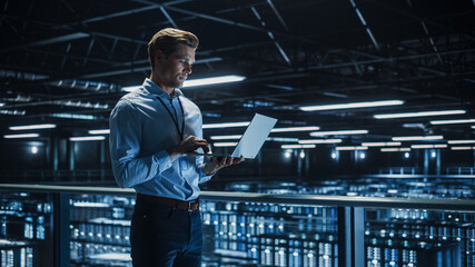 Handsome Smiling IT Specialist Using Laptop Computer in Data Center. Succesful Businessman and e-Business Entrepreneur Overlooking Server Farm Cloud Computing Facility.