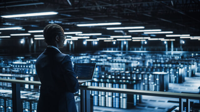 Female IT Specialist Using Laptop Computer in Data Center, Walking on a Bridge Overlooking Big Server Farm Cloud Computing Facility. Businesswoman, e-Business Entrepreneur.