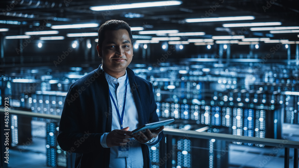 Poster Handsome Smiling IT Specialist Using Tablet Computer in Data Center, Looking at Camera. Succesful Businessman and e-Business Entrepreneur Overlooking Server Farm Cloud Computing Facility.