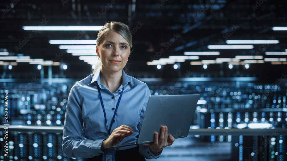 Poster Portrait of Beautiful Smiling IT Specialist Using Laptop Computer in Data Center, Looking at Camera. Succesful Female e-Business Engineer Working in Big Server Farm Cloud Computing Facility.