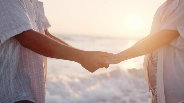 Unrecognizable couple holding hands on seashore at sunset close up