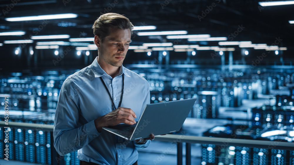 Poster Handsome Smiling IT Specialist Using Laptop Computer in Data Center. Succesful Businessman and e-Business Entrepreneur Overlooking Server Farm Cloud Computing Facility.