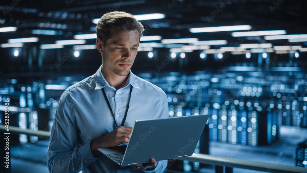 Poster Handsome Smiling IT Specialist Using Laptop Computer in Data Center. Succesful Businessman and e-Business Entrepreneur Overlooking Server Farm Cloud Computing Facility.
