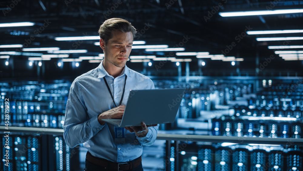 Poster Handsome Smiling IT Specialist Using Laptop Computer in Data Center. Succesful Businessman and e-Business Entrepreneur Overlooking Server Farm Cloud Computing Facility.