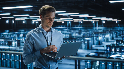 Handsome Smiling IT Specialist Using Laptop Computer in Data Center. Succesful Businessman and e-Business Entrepreneur Overlooking Server Farm Cloud Computing Facility.