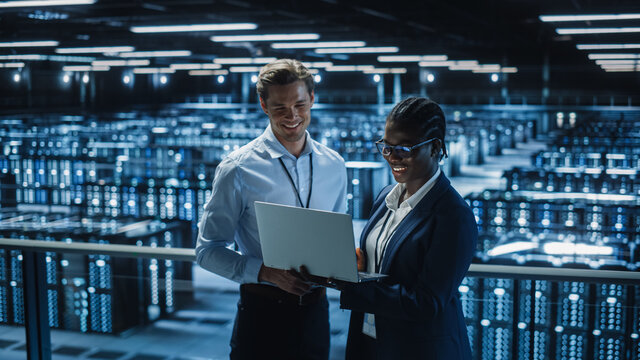 Data Center Female System Administrator And Male IT Specialist Talk, Use Laptop. Information Technology Engineers Work On Cyber Security Protection In Cloud Computing Server Farm.