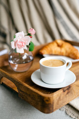 Fresh coffee with croissant on a wooden tray with a pink tulip in a glass vase.