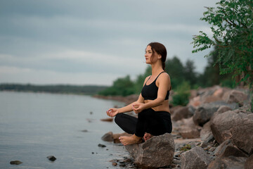 Fototapeta na wymiar Woman in black sport suit is doing meditation, yoga pose (asana), work out outside near beautiful river and beautiful landscape with wild nature
