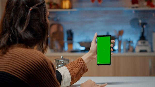 Caucasian woman holding smartphone with green screen display in kitchen with tree, decorations and ornaments. Young person using telephone gadget while preparing for christmas eve