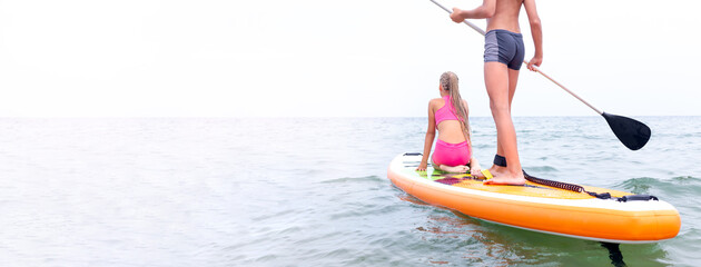 Two persons, boy and girl, on the paddle board on the calm sea surface moving forward to the horizon. SUP boarding, water sports. Active healthy lifestyle. Wide banner with copy space.