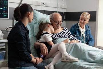 Obraz premium Caring granddaughter hugging elderly old grandfather supporting him after medical surgery in hospital ward. Senior man resting in bed recovering during clinical therapy. Healthcare support