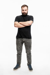 Naklejka premium A man with a beard is standing, arms crossed over his chest. Young man in jeans and a black T-shirt. Full height. White background. Vertical.