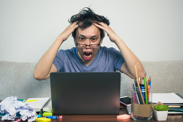 Crazy shouting man with messy hair, wide open mouth and holding hands on head sitting at home table using laptop. Work from home