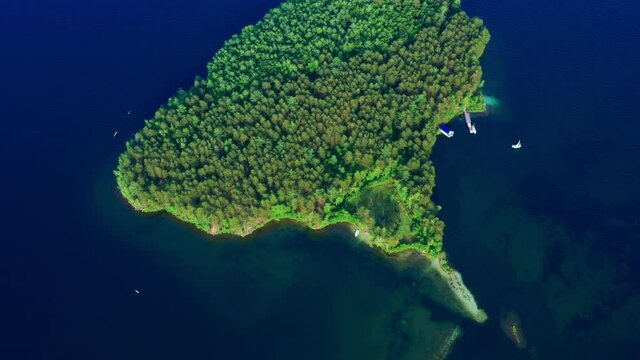 Small beautiful island in ocean covered with green plants, trees taken by drone. Aerial, top view of river ait, eyot, white yachts, seagulls flying over it. Sea isle, blue water surface