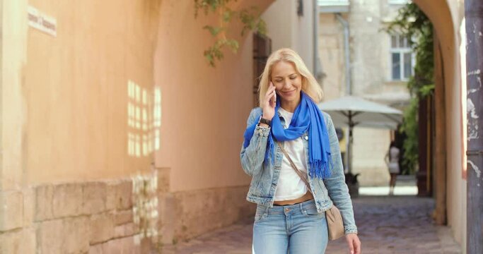 Portrait of Caucasian young pretty woman speaking on smartphone while walking on street in town with smile on face. Beautiful blonde female chatting on mobile phone call outdoors, conversation concept