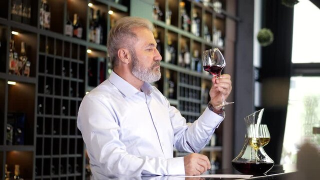 An elderly, bearded, gray-haired, well-dressed sommelier, a male winemaker checks the quality of wine. He writes on paper, describes the taste of wine. The senior sommelier is tasting the wine.
