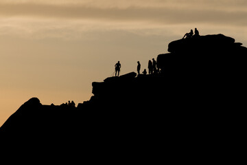 Silhouette of lots of people watching a climber at the summit of stanage edge. Climber wears a harness with tools like nuts, ropes and carabiners.