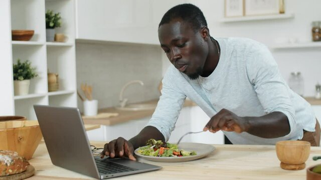 Young Afro-American man leaning on kitchen table, eating salad and surfing the Internet on laptop