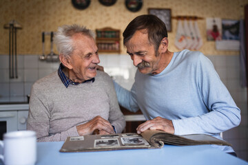 Portrait of man with elderly father sitting at the table indoors at home, looking at photo album.