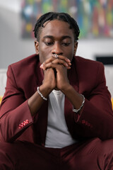 Dark skinned man with dreadlocked wearing smart suit sitting in office hands resting on knees joined in front of face worry sadness pondering company problems money troubles thinking of solution