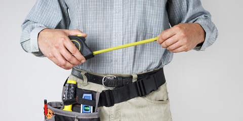Builder's hands hold yardstick for measuring while working and tools in waist bag, close-up on light background
