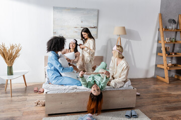 group of joyful interracial friends in pajamas sitting on bed during slumber party