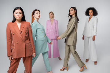 full length of interracial models in stylish suits walking on grey