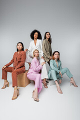 full length of interracial models in stylish pastel suits sitting while posing on grey