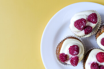 Cupcakes with white cream and raspberries in a white plate on a yellow background. Sweet homemade cakes close-up on a colored background.
