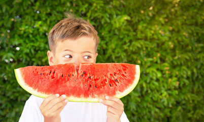 Happy child (boy) is eating a red juicy watermelon. Caucasian kid smiling and having fun. Concept of healthy food, happy childhood, summer vacation. Nature background. Copy space.