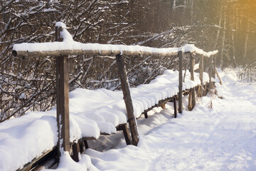Old wooden footbridge over a stream under the snow in a winter park with sun rays