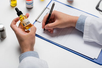 partial view of doctor with vial of cbd oil writing prescription on clipboard