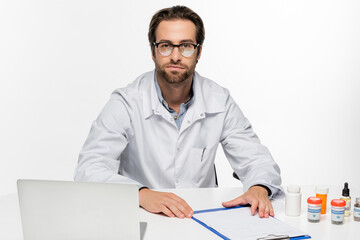 physician looking at camera near containers with medical hemp, laptop and clipboard isolated on white