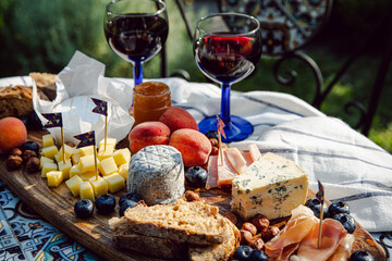 Cheese platter with different cow and goat cheeses, meat, nuts, Summer fruits and berries, jam and rye bread on wooden board.with red wine on little rustic table in the garden.
