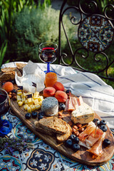 Cheese platter with different cow and goat cheeses, meat, nuts, Summer fruits and berries, jam and rye bread on wooden board.with red wine on little rustic table in the garden.
