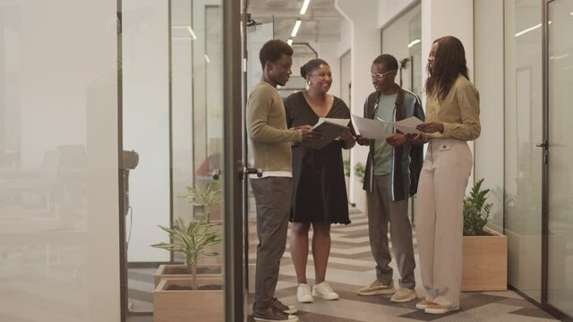 Full-length shot of African American group of colleagues holding business documents discussing work standing in modern office