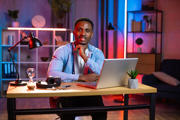 Fototapeta na wymiar Evening leisure or remote work concept. Good-looking smiling black-skinned man, sitting in front of laptop at home and posing to camera. Colorful nigh lights on the background